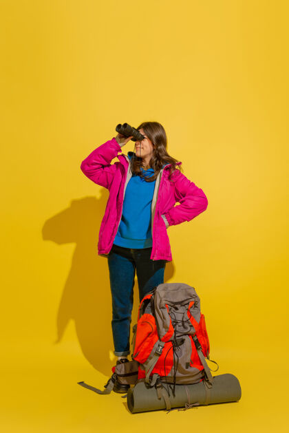 指向黄色摄影棚墙上 一个带着包和望远镜的快乐的年轻旅游女孩的画像快乐手手提箱