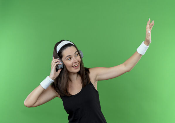 年轻戴着耳机戴着头带的年轻健身女士面带微笑 站在绿色的墙壁上挥手致意手头带微笑