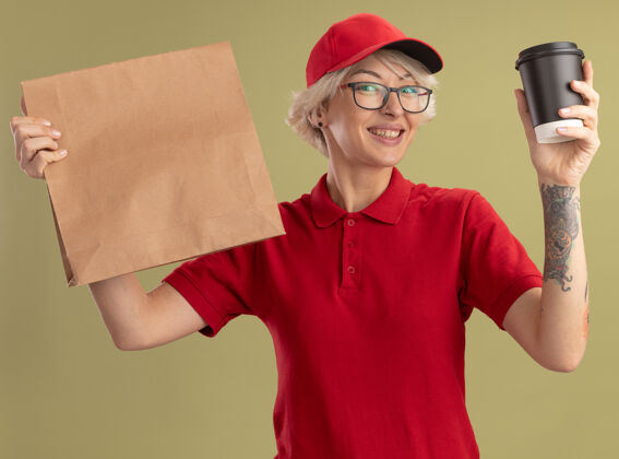 包装身穿红色制服 戴着帽子 戴着眼镜 手里拿着纸包和咖啡杯的年轻送货员站在绿色的墙上 脸上洋溢着幸福的笑容眼镜年轻杯子