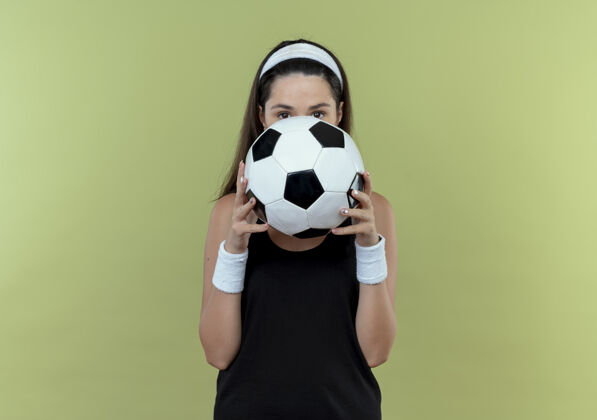 年轻戴着足球头巾的年轻健身女士把脸藏在球后 站在轻质墙上偷看轻女子球