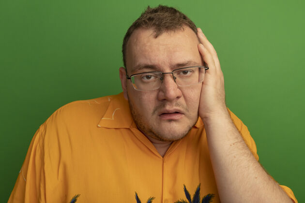 手戴着眼镜 穿着橙色衬衫的男人站在绿色的墙上 手放在头上 又烦又累烦头人