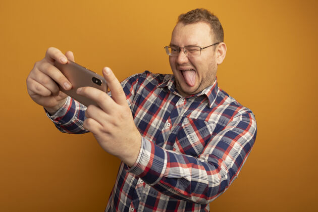 站立戴着眼镜和格子衬衫的男人用智能手机自拍微笑着伸出舌头站在橙色的墙上舌头检查微笑