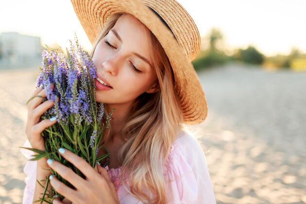 肖像戴着草帽的温柔漂亮的女人在海边阳光明媚的海滩上摆着一束鲜花特写肖像欢呼皮肤护理帽子