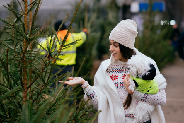 一起市场上一棵绿色的圣诞树旁 一个抱着一只白色狗的女人快乐可爱圣诞节