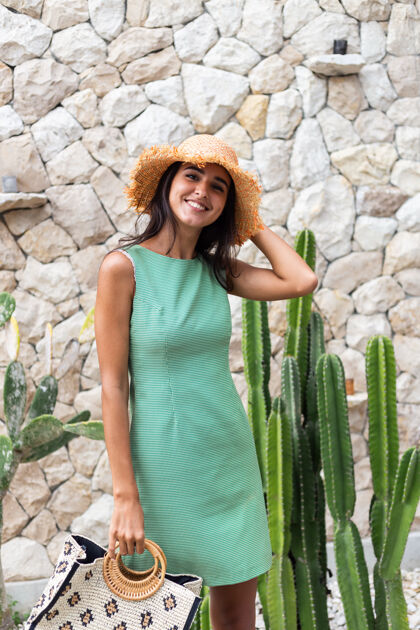 配件在白色石墙和仙人掌的背景下 穿着优雅的夏日绿色连衣裙 手拿袋子 头戴草帽的时尚快乐可爱微笑女人的肖像帽子成人女士