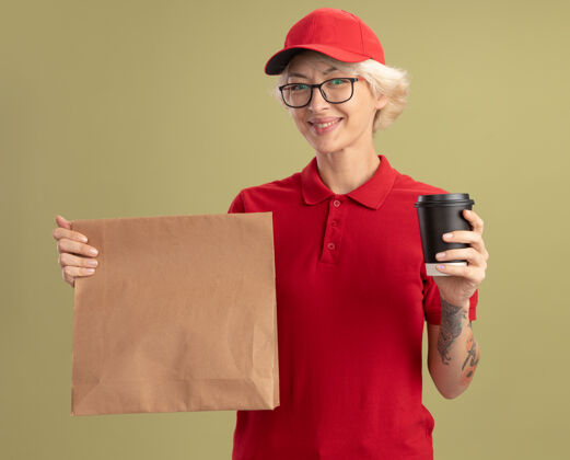 包装身着红色制服 戴着眼镜 手持纸袋和纸杯的年轻送货员友好地站在绿色的墙上朋友送货年轻