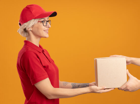 眼镜身着红色制服 戴着眼镜的年轻送货员站在橙色的墙上 友好地微笑着给你的顾客送纸盒顾客微笑赠送