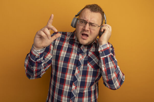手指情绪激动的男人戴着眼镜 穿着格子衬衫 戴着耳机 看着一旁 食指站在橙色的墙上表演情绪目录