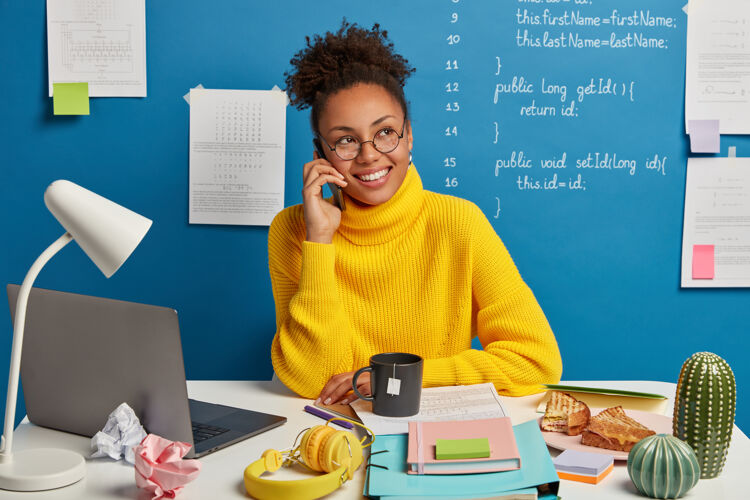 非洲裔积极的黑皮肤女孩打电话 讨论改善和发展的商业项目 穿着黄色毛衣 看一边 在蓝色背景下摆姿势极客报告通话