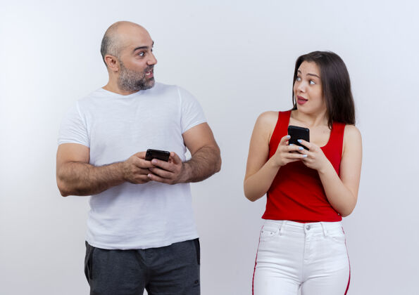 两者令人印象深刻的成年夫妇都拿着手机 看着对方隔离在白墙上情侣抱着手机