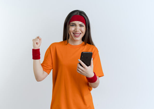 年轻快乐的年轻运动女性戴着头带和手环拿着手机做着“是”的手势 隔离在白色墙壁上 留着复印空间头带戴手机