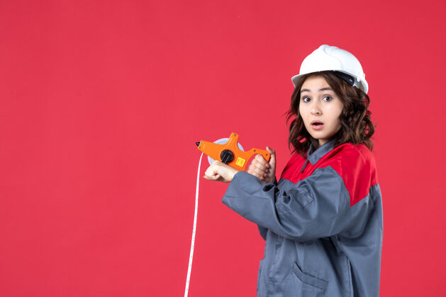 磁带穿着制服 戴着安全帽 在红墙上用卷尺测量的惊讶女建筑师的特写镜头漂亮测量硬