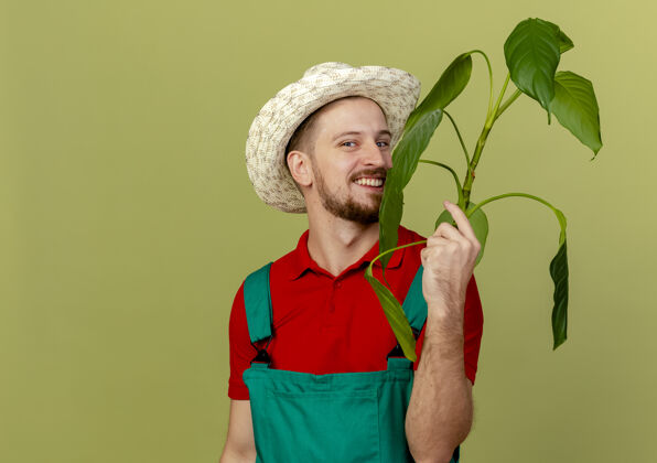 制服快乐的年轻英俊的斯拉夫园丁在制服和帽子举行植物隔离在橄榄绿的墙壁与复制空间年轻英俊植物