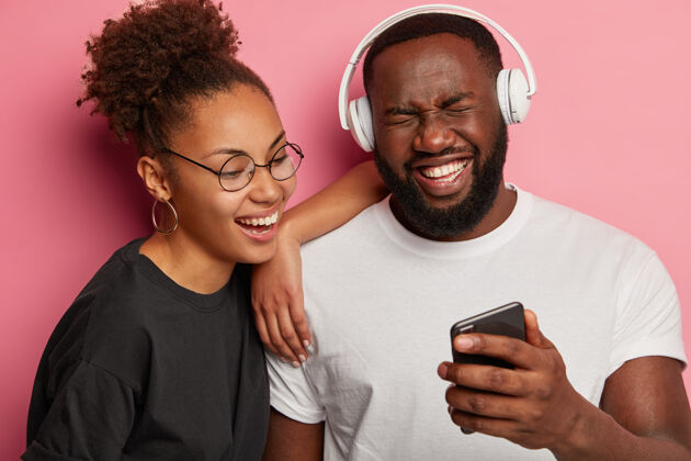 情侣喜出望外的黑人潮人拿着智能手机 和女友一起在智能手机上看搞笑电影时开心地笑着 用立体声耳机卷发快乐朋友