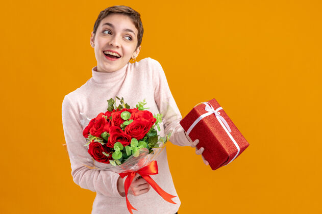 欢呼一个留着短发的年轻女子 手里拿着一束红玫瑰 手里拿着一件礼物 看上去很快乐 站在橘色的墙上微笑着情人节的概念期待举行玫瑰
