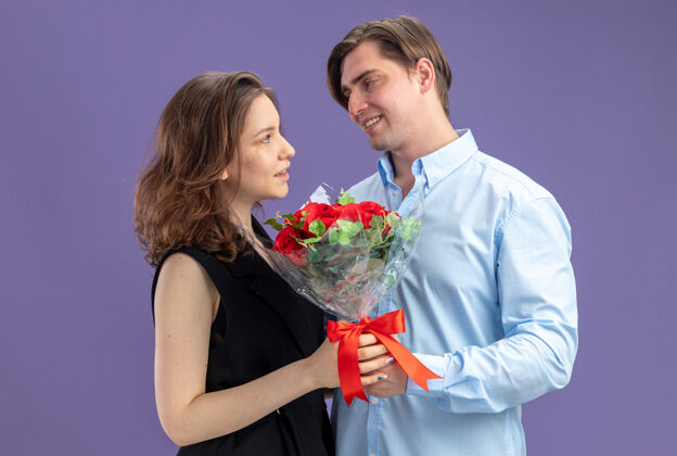 年轻年轻漂亮的情侣幸福的男人站在蓝色的墙上 送一束红玫瑰给他微笑可爱的女友庆祝情人节女朋友情侣男人