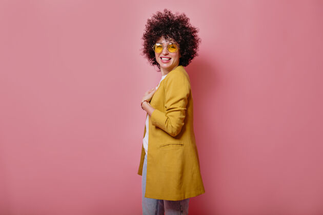 非洲满意的年轻微笑的女孩卷发穿着黄色夹克看着前面粉红色的墙治疗皮肤护理大笑