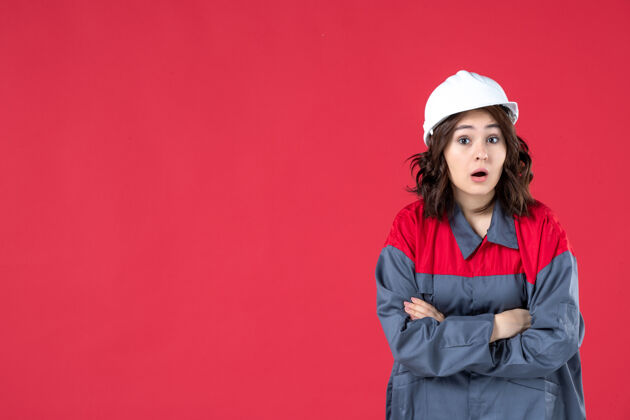手臂正面近距离观看困惑的女建筑工人穿着制服 戴着安全帽 双臂交叉在隔离的红墙上制服肖像前面