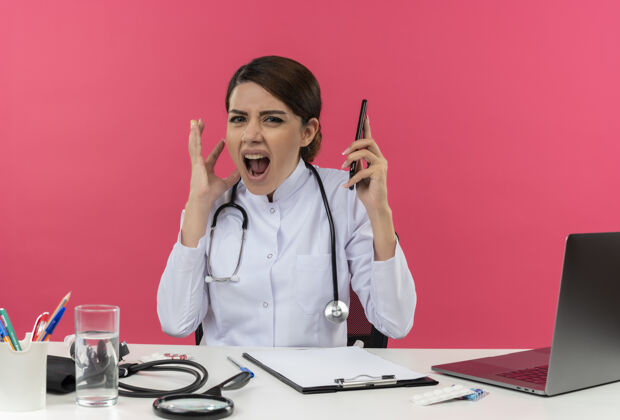 听诊器紧张的年轻女医生穿着医用长袍和听诊器坐在办公桌旁 手里拿着医疗工具和笔记本电脑 手里拿着手机 手举在空中 在粉红色的墙上尖叫着空气保持穿