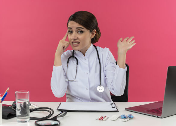 年轻不知所措的年轻女医生穿着医用长袍 听诊器坐在办公桌旁 手里拿着医疗工具和笔记本电脑 手指指着头 粉红色的墙上只露出一只空手头显示手