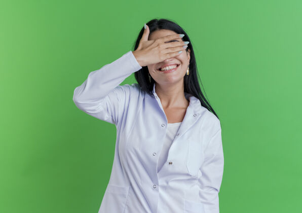 微笑面带微笑的年轻女医生 穿着医用长袍 用手遮住眼睛 隔离在绿色墙壁上 留有复印空间医疗穿年轻