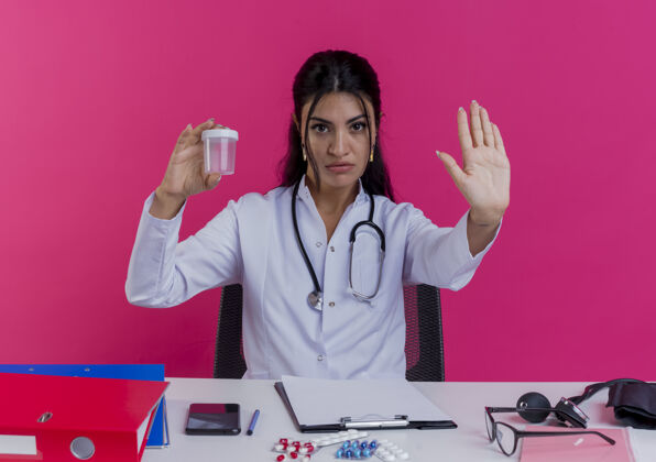 手势年轻严格的女医生穿着医用长袍和听诊器坐在办公桌前拿着医疗工具拿着医用烧杯做停止手势隔离在粉红色的墙上烧杯医生穿