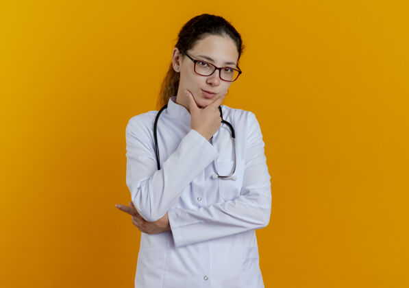 医学想着年轻的女医生 穿着医用长袍 戴着眼镜的听诊器 把下巴孤立在橘黄色的墙上穿年轻长袍