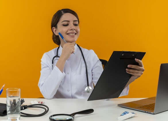坐着面带微笑的年轻女医生穿着医用长袍和听诊器坐在办公桌旁 拿着医疗工具和笔记本电脑 看着剪贴板 触摸着隔离在黄色墙上的钢笔的脸听诊器长袍笔记本电脑