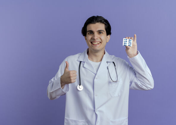 拇指面带微笑的年轻男性医生 穿着医用长袍 听诊器 展示一包胶囊 拇指朝上 隔离在紫色的墙上 留有复印空间男性微笑医生