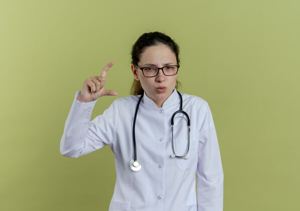 长袍自信的年轻女医生穿着医用长袍 戴着听诊器和眼镜 在橄榄绿的墙上显示出孤立的大小年轻自信听诊器