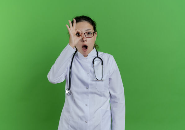 表情令人印象深刻的年轻女医生穿着医用长袍和听诊器 戴着眼镜 在绿色的墙上做着孤立的看手势 还有复印空间手势穿着听诊器