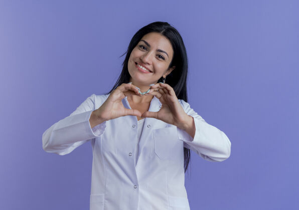 医生面带微笑的年轻女医生穿着医用长袍 在紫色的墙上做着孤立的心牌 还有复制空间心脏微笑穿着