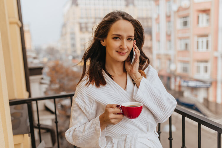成人绿眼睛 黑发 穿着白袍的女人在阳台上打电话女孩看着镜头 端着一杯红茶休息健康脸