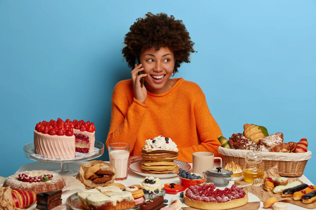 女性快乐的非洲发型女士通过手机进行愉快的交谈 喜欢吃美味的蛋糕 喜欢吃奶油煎饼 喜欢吃甜食 被隔离在蓝色的墙壁上积极女人咖啡