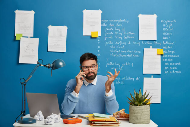 专业年轻的男企业家在电话交谈中思考商业解决方案 困惑地举手 拿着笔记本 皱巴巴的纸和笔记本电脑坐在白色的办公桌旁灯困惑男性
