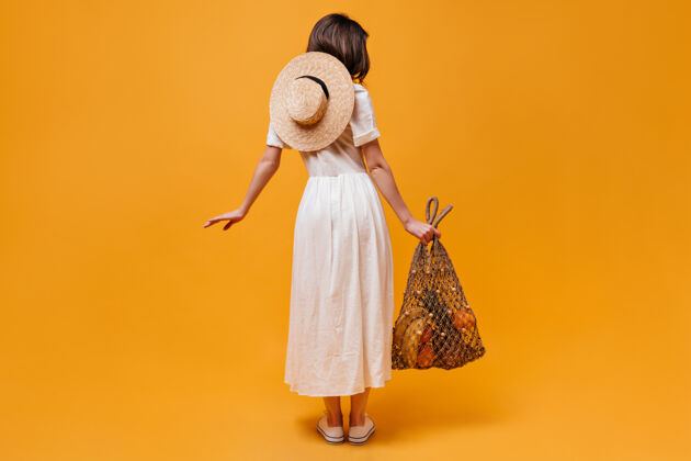 脸身着迷笛裙 头戴草帽 背着橘色背景的水果串袋的女孩的全长照片卷发长波浪发