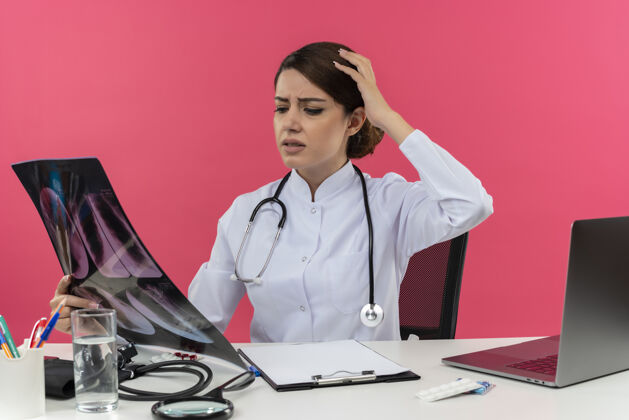 长袍失望的年轻女医生穿着医用长袍和听诊器坐在办公桌旁 拿着医疗工具和笔记本电脑 看着x光照片摸着头粉色看坐着