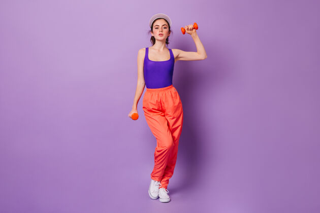 深色穿着紫色运动上衣和橙色裤子的女士用红色哑铃练习双手80年代身体头发