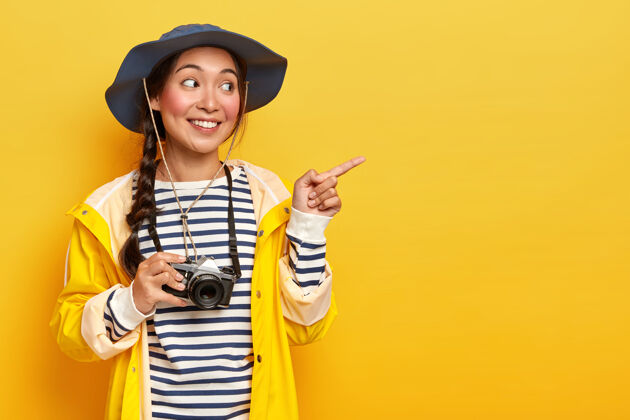 摄影师满意的韩国女人戴着头饰 条纹套头衫和黄色雨衣 把食指指放在一边 宣传复制空间 携带复古相机 在野外旅行 有一次惊险的探险复古成人旅行者