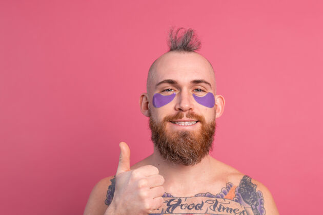 强壮欧洲帅哥留着胡子纹身上身的男人戴着紫色眼罩对着镜头摆粉红色的姿势治疗健康抗衰老