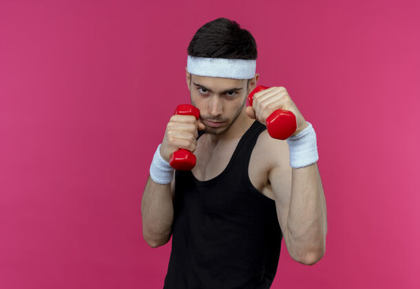 站立年轻的运动型男子戴着头巾 戴着哑铃 紧张而自信地在粉色的头上锻炼头带男人自信