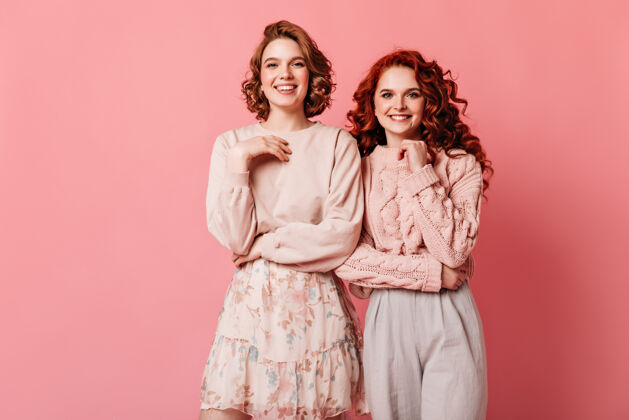 姐妹摄影棚拍摄的迷人的女孩孤立在粉红色的背景两个时尚的朋友微笑着看着相机卷曲微笑孤立的粉色