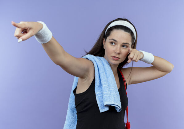 头带戴着头巾 肩上扛着毛巾的年轻健身女士看起来自信满满 手指和双手指向蓝色墙壁的一侧健身手站