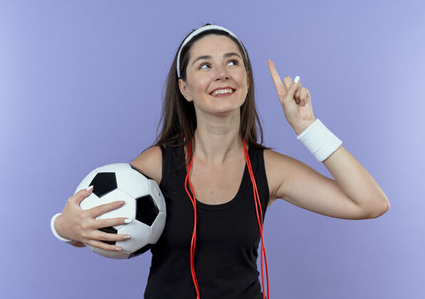 头带戴着头巾 脖子上系着跳绳 举着足球 手指朝上 面带微笑的年轻健身女士站在蓝色的墙上 有了新的想法手指抱着绳子