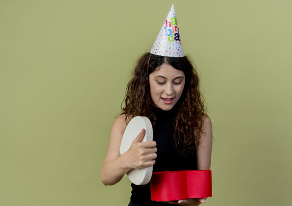 盒子年轻漂亮的女人卷发戴着节日帽拿着礼盒看着它惊喜而快乐的生日派对概念过了光看卷发生日