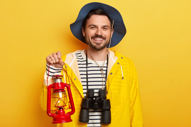 休闲英俊的没刮胡子的男人拿着煤油灯 望远镜 准备远征或旅行 戴着帽子和雨衣 隔离在黄色的墙壁上室内阳刚帽子