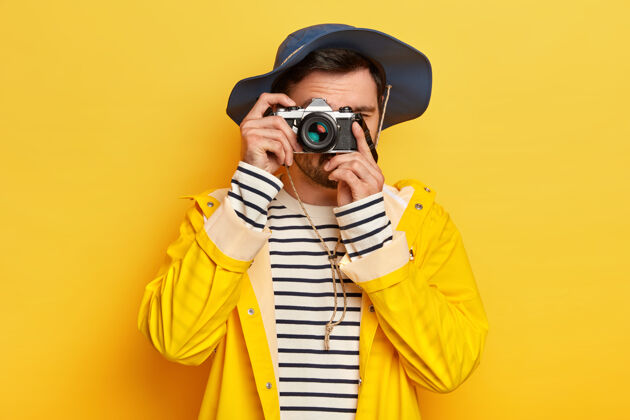 防水年轻活跃的男性旅行者带着复古相机拍照 戴着帽子 穿着雨衣在雨天旅行 对着黄色的墙壁摆姿势散步摄影脸