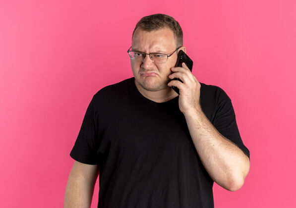 黑色戴眼镜的超重男子身穿黑色t恤 一边用粉色手机交谈一边显得不高兴而手机穿