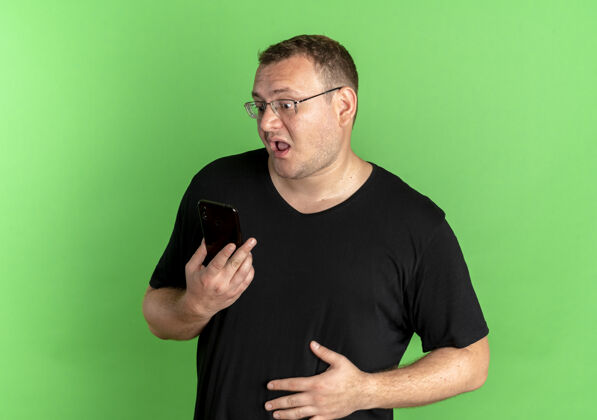 惊喜一个戴眼镜 身穿黑色t恤的超重男子看着自己的智能手机屏幕 对绿色感到困惑和惊讶男士T恤表情
