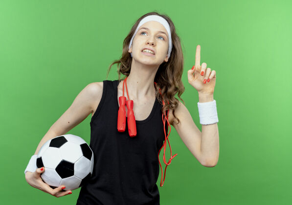 抱着穿着黑色运动服的年轻健身女孩 头上戴着头带 脖子上系着跳绳 手里拿着足球 抬起头来 食指对着绿色微笑健身黑色向上